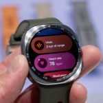 Photo officielle de la Samsung Galaxy Watch 9 tenue en main, écran affichant les widgets santé (vitals, fréquence cardiaque, tension)
